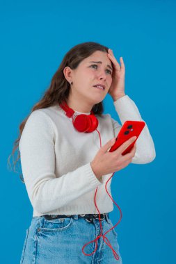 Beyaz kazaklı, elinde kulaklıkla kırmızı akıllı telefon tutan endişeli genç bir kadın, ekranı, alnı mavi arka planı izlerken şok ve stres içinde tepki veriyor.