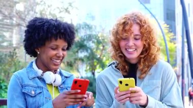Güneşli bir park bankında sarı ve kırmızı telefonları karıştıran iki çok ırklı arkadaş viral videoları, sosyal medya güncellemelerini ve birlikte neşeli dijital yaşam tarzı anlarını paylaşıyorlar.. 