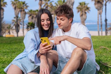 Palmiye ağaçlarının altında deniz kenarında oturan genç çift, birlikte sarı bir akıllı telefona bakarken gülümsüyor, ekrana işaret eden erkek arkadaş, aşkı, seyahat anlarını ve bugün dijital bağlanmayı yakalıyor. 