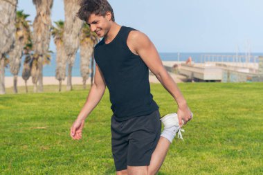 Güneşli sahil çimenliği, siyah tanklı zinde bir genç adam ve şort, kaslarını çalıştırmak için bir ayağını kaslarına doğru uzatarak palmiyeler ve mavi sular arasında dinamik bir egzersiz yapıyor. 