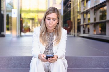 Beyaz takım elbiseli iş kadını modern bina basamaklarında oturuyor, akıllı telefona mesaj atmaya odaklanıyor, iş molası, iletişim, teknoloji, kentsel yaşam tarzı ve profesyonel açık hava düzeni kavramını gösteriyor 