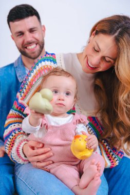 Oturma odasındaki kanepede gülümseyen ebeveynler pembe tulumlu kızlarını sarı ördek ve yeşil kaplumbağa oyuncaklarıyla eğlendirip neşeli aile bağlarını ve erken çocukluk gelişimini kapalı alanda yakalıyorlar.