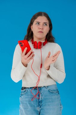 Beyaz kazaklı, elinde kulaklıkla kırmızı akıllı telefon tutan endişeli genç bir kadın, ekranı, alnı mavi arka planı izlerken şok ve stres içinde tepki veriyor.