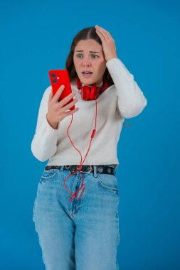 Beyaz kazaklı, elinde kulaklıkla kırmızı akıllı telefon tutan endişeli genç bir kadın, ekranı, alnı mavi arka planı izlerken şok ve stres içinde tepki veriyor.