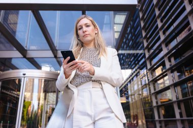 Akıllı telefon, modern cam bina, kentsel şehir, alçak açı, kariyer, profesyonel, teknoloji, dijital iletişim, çalışma, liderlik, başarı gibi konularda kendine güvenen beyaz takım elbiseli bir iş kadını. 