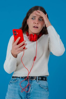 Beyaz kazaklı, elinde kulaklıkla kırmızı akıllı telefon tutan endişeli genç bir kadın, ekranı, alnı mavi arka planı izlerken şok ve stres içinde tepki veriyor.
