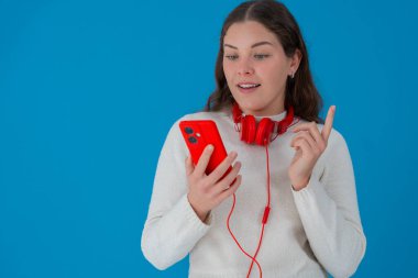 Kırmızı kulaklık takan, kırmızı kulaklık takan, telefonuna bakarken işaret parmağını kaldıran, gülümseyen genç bir kadın. 