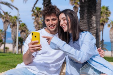 Genç bir adam ve kadın, deniz kenarındaki palmiye ağaçlarının altındaki ahşap bankta oturuyor. Sarı akıllı telefona birlikte bakıp, sosyal medyanın ve açık hava rahatlığının keyfini çıkarırken gülümsüyorlar. 