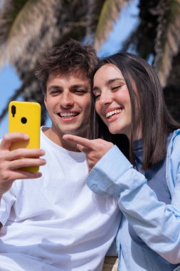 Genç bir adam ve kadın, deniz kenarındaki palmiye ağaçlarının altındaki ahşap bir bankta oturuyor, birlikte sarı akıllı telefona bakarken gülümsüyorlar, sosyal medyanın ve açık hava rahatlığının keyfini çıkarıyorlar. Dikey