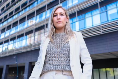 Hafif bir takım elbise giyen, ciddi şekilde kameraya bakan, modern bir ofis binasının önünde duran, liderliği, kararlılığı ve profesyonelliği temsil eden kendine güvenen bir iş kadını.. 