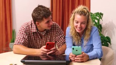 Mutlu çift masada oturuyor, gülümsüyor ve birlikte akıllı telefonlara bakıyor, sosyal medyaya ve mesajlara bakıyorlar, masadaki dizüstü bilgisayara, rahatlamış ruh hali, modern yaşam tarzı, iletişim, teknoloji. 
