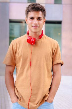 Boynuna kırmızı kulaklıklar takmış, modern mimari ve güneş ışığını yansıtan şehir ortamında duran kameraya içten bir şekilde gülümseyen genç bir adamın portresi. 