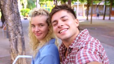 Mutlu genç çift güneşli bir günün tadını çıkarıyor, birlikte oturup kameraya bakarken sıcacık gülümsüyor, neşeli ve rahat bir anı ve doğal şehir parkı ortamını yakalıyorlar. 