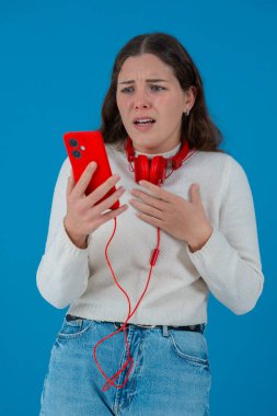 Beyaz kazaklı, elinde kulaklıkla kırmızı akıllı telefon tutan endişeli genç bir kadın, ekranı, alnı mavi arka planı izlerken şok ve stres içinde tepki veriyor.