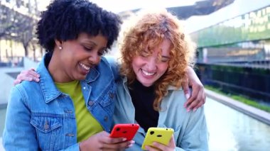 Açık havada yan yana duran mutlu genç kadınlar, renkli akıllı telefonlar tutuyorlar ve ekranlara bakarken gülümsüyorlar, sosyal medyadan hoşlanıyorlar ya da kentsel bir ortamda su özelliğinin yanında eğlenceli bir video izliyorlar. 