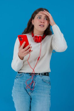 Beyaz kazaklı, elinde kulaklıkla kırmızı akıllı telefon tutan endişeli genç bir kadın, ekranı, alnı mavi arka planı izlerken şok ve stres içinde tepki veriyor.