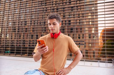 Boynuna kırmızı kulaklık takan sıradan bir genç, arka planda binalar olan modern bir şehir ortamında dikilirken akıllı telefonuna bakıyor. 