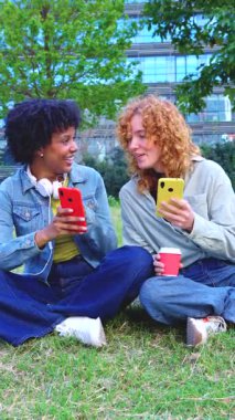 Şehir parkında çimenlerde oturan mutlu genç kadınlar, ellerinde akıllı telefonlar ve kahve fincanı, birlikte vakit geçiriyorlar ve içeriklerini paylaşıyorlar ve boyunlarına günlük giysiler ve kulaklıklar takıyorlar. 