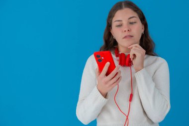 Kırmızı akıllı telefon ve kulaklık tutan beyaz kazaklı genç bir kadın, elleri çenesinde düşünceye bakıyor, mavi arka planda izole edilmiş, merak ya da şüphe gösteriyor. Boşluğu kopyala