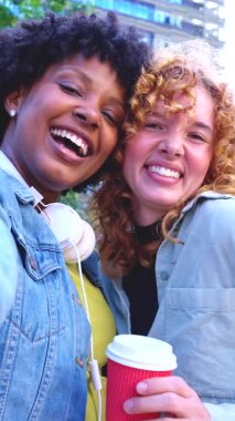 Açık havada gülümseyen iki genç kadının dikey selfie 'si, günlük ceketler giyiyor, biri paket kahve bardağı tutuyor, binalarla dolu bir şehir ortamında birlikte vakit geçiriyorlar.. 