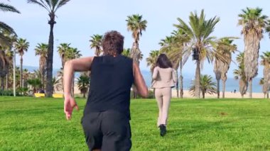 Arka planda gülümseyen genç bir çift palmiyeli bir kıyı yolunda yan yana koşuyor, açık hava egzersizinden, spor için mükemmel güneşli ve manzaralı bir ortamda sağlıklı bir yaşam sürüyor.. 