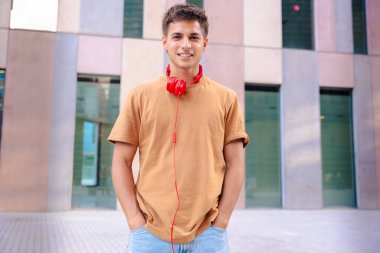 Boynuna kırmızı kulaklıklar takmış, modern mimari ve güneş ışığını yansıtan şehir ortamında duran kameraya içten bir şekilde gülümseyen genç bir adamın portresi..