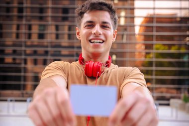 Boynunda kırmızı kulaklıklarla gülümseyen genç bir adam açık havada kameraya boş bir kartvizit gösteriyor. Metal parmaklıklar ve arkadaki bulanık binalarla modern bir şehir arka planının önünde duruyor. 