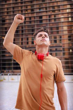 Heyecanlı genç adam, kutlama için gülümsüyor ve boynuna kırmızı kulaklıklar takarak yumruğunu kaldırıyor, şehir dışında modern binalar ve sıcak günışığıyla dikey bir ortamda duruyor.