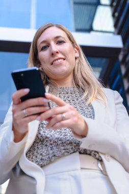 Akıllı telefon, modern cam bina, kentsel şehir, alçak açı, kariyer, profesyonel, teknoloji, dijital iletişim, çalışma, liderlik, başarı gibi konularda kendine güvenen beyaz takım elbiseli bir iş kadını. 