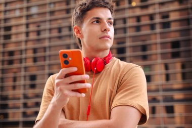 Boynuna kırmızı kulaklık takan sıradan bir genç, arka planda binalar olan modern bir şehir ortamında dikilirken akıllı telefonuna bakıyor. 
