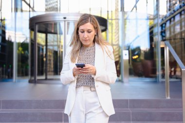 Akıllı telefon, modern cam bina, kentsel şehir, alçak açı, kariyer, profesyonel, teknoloji, dijital iletişim, çalışma, liderlik, başarı gibi konularda kendine güvenen beyaz takım elbiseli bir iş kadını. 