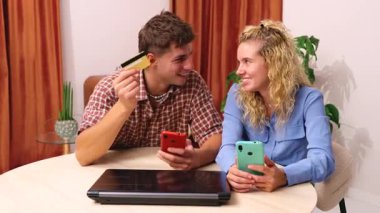 Akıllı telefonlar kullanarak bir masada oturan ve ellerinde online alışveriş yapmak için bir kredi kartı tutan genç bir çift, önlerinde bir dizüstü bilgisayarla, rahat bir ev ortamında e-ticaret alışverişinin keyfini çıkarıyorlar.