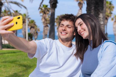 Mutlu genç çift, güneşli bir sahil gezintisinde eğlenceli bir selfie için poz veriyor, o parlak sarı bir akıllı telefon tutarken, o da yaz havasını yakalıyor, seyahat anılarını, aşkı yakalıyor.. 