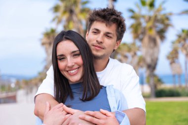 Genç çift, palmiyeli bir sahil geçidinde kucaklaşıyor, kız arkadaş gibi gülüyor, kollarını arkadan sarıyor, neşeli bir aşk yakalıyor, seyahat titreşimleri alıyor, yaz yaşam tarzı, bağlantı. 