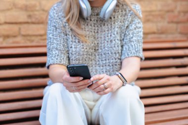 Kulaklık takan tanınmayan bir kadın sıradan kıyafetlerle dışarıda duruyor, ciddi değil ve elinde akıllı bir telefon, şehir parkı ayarlarıyla kameraya bakıyor..