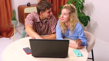 Akıllı telefonlar kullanarak bir masada oturan ve ellerinde online alışveriş yapmak için bir kredi kartı tutan genç bir çift, önlerinde bir dizüstü bilgisayarla, rahat bir ev ortamında e-ticaret alışverişinin keyfini çıkarıyorlar.