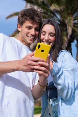 Palmiye ağaçlarının altında deniz kenarında oturan genç çift, birlikte sarı bir akıllı telefona bakarken gülümsüyor, ekrana işaret eden erkek arkadaş, aşkı, seyahat anlarını ve bugün dijital bağlanmayı yakalıyor. 