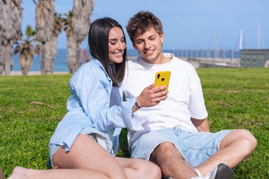 Palmiye ağaçlarının altında deniz kenarında oturan genç çift, birlikte sarı bir akıllı telefona bakarken gülümsüyor, ekrana işaret eden erkek arkadaş, aşkı, seyahat anlarını ve bugün dijital bağlanmayı yakalıyor. 