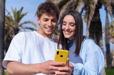 Palmiye ağaçlarının altında deniz kenarında oturan genç çift, birlikte sarı bir akıllı telefona bakarken gülümsüyor, ekrana işaret eden erkek arkadaş, aşkı, seyahat anlarını ve bugün dijital bağlanmayı yakalıyor. 