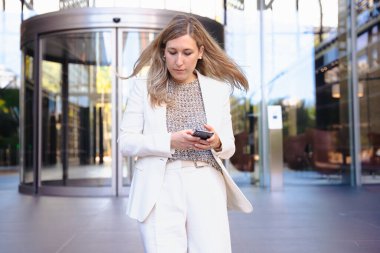 Akıllı telefon, modern cam bina, kentsel şehir, alçak açı, kariyer, profesyonel, teknoloji, dijital iletişim, çalışma, liderlik, başarı gibi konularda kendine güvenen beyaz takım elbiseli bir iş kadını. 