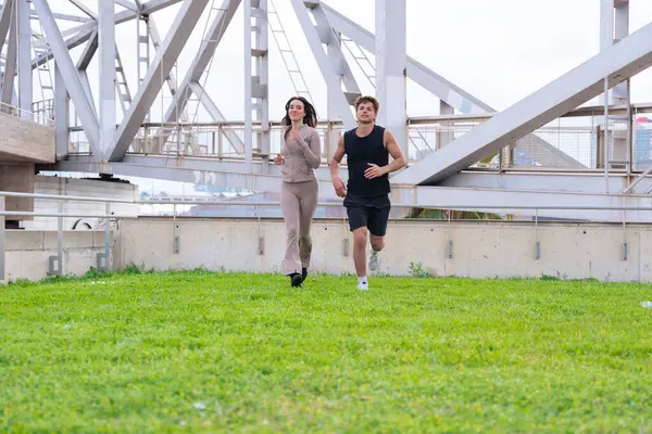Genç bir adam ve kadın açık havada yeşil çimlerin üzerinde koşuyorlar, büyük metal bir köprünün altında, spor giyiyorlar, spor kıyafetleri giyiyorlar, fitness aktivitelerinin keyfini çıkarıyorlar, sağlıklı bir yaşam tarzı, bulutlu bir günde şehirde egzersiz yapıyorlar. 