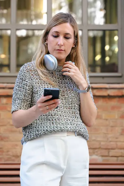 Boynuna kulaklık takmış genç sarışın bir kadın akıllı telefonuna bakarken gülümsüyor. Günlük kıyafet, şehir parkı dekorasyonu, arka planda tuğla duvar. Dikey.