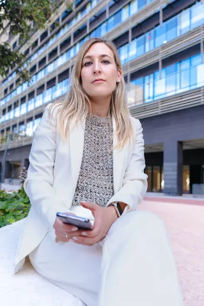 Akıllı telefon, modern cam bina, kentsel şehir, alçak açı, kariyer, profesyonel, teknoloji, dijital iletişim, çalışma, liderlik, başarı gibi konularda kendine güvenen beyaz takım elbiseli bir iş kadını. 