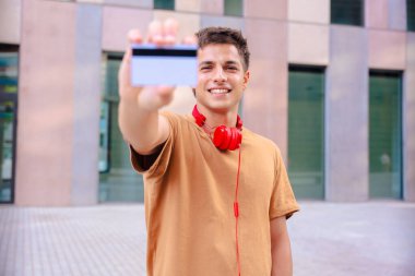 Boynunda kırmızı kulaklıklarla gülümseyen genç bir adam açık havada kameraya boş bir kartvizit gösteriyor. Metal parmaklıklar ve arkadaki bulanık binalarla modern bir şehir arka planının önünde duruyor. 