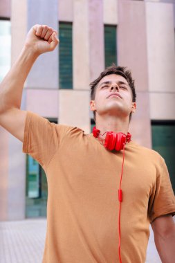 Heyecanlı genç adam, kutlama için gülümsüyor ve boynuna kırmızı kulaklıklar takarak yumruğunu kaldırıyor, şehir dışında modern binalar ve sıcak günışığıyla dikey bir ortamda duruyor.