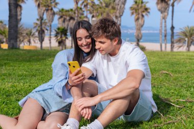Palmiye ağaçlarının altında deniz kenarında oturan genç çift, birlikte sarı bir akıllı telefona bakarken gülümsüyor, ekrana işaret eden erkek arkadaş, aşkı, seyahat anlarını ve bugün dijital bağlanmayı yakalıyor. 