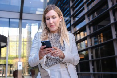 Akıllı telefon, modern cam bina, kentsel şehir, alçak açı, kariyer, profesyonel, teknoloji, dijital iletişim, çalışma, liderlik, başarı gibi konularda kendine güvenen beyaz takım elbiseli bir iş kadını. 