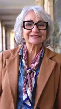 Şık gözlüklü, gri saçlı, şık giyimli, şehir ortamında ayakta duran, kendine güveni, sıcaklığı ve doğal ışıkta zamansız güzelliğiyle gülümseyen son sınıf öğrencisinin dikey portresi. 