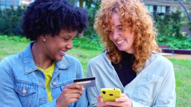  Neşeli Afrikalı Amerikalı ve Kafkasyalı arkadaşlar ellerinde renkli akıllı telefonlar ve bir kredi kartı ile bir şehir parkında duruyorlar. Nakitsiz kolaylığı kutluyorlar, alışveriş başarılarını paylaşıyorlar ve dijital bir yaşam tarzları var.