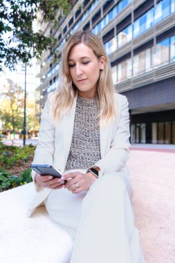 Akıllı telefon, modern cam bina, kentsel şehir, alçak açı, kariyer, profesyonel, teknoloji, dijital iletişim, çalışma, liderlik, başarı gibi konularda kendine güvenen beyaz takım elbiseli bir iş kadını. 